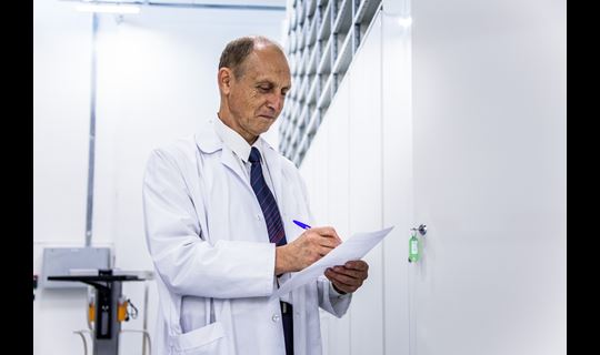 A man in a lab coat writes notes on a piece of paper standing in a pharmaceutical storage depot.