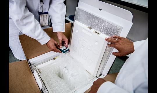 Two people in lab coats take a reading from a digital monitor inside an insulated package.