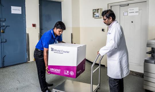 A World Courier driver loads a large box onto a cart pushed by a man in a lab coat.